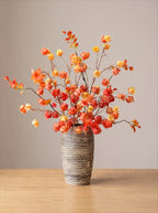 Autumn artificial flower branch arrangement in a ribbed gray ceramic vase, set on a light wood tabletop against a neutral beige background.