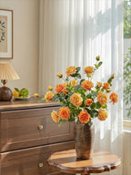 A bouquet of orange and yellow roses with green leaves arranged in a ribbed brown ceramic vase, placed on a small wooden table in a bright room with sheer white curtains, a wooden dresser, and a lamp in the background.