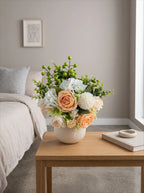 A floral arrangement in a round white vase sits on a light wood table in a softly lit bedroom. The bouquet includes peach roses, white dahlia-like blooms, green berries, and green foliage.
