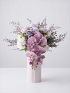A floral arrangement featuring pink and purple orchids, lavender-toned roses, white blossoms, and green foliage in a ribbed white ceramic vase against a light gray studio background.