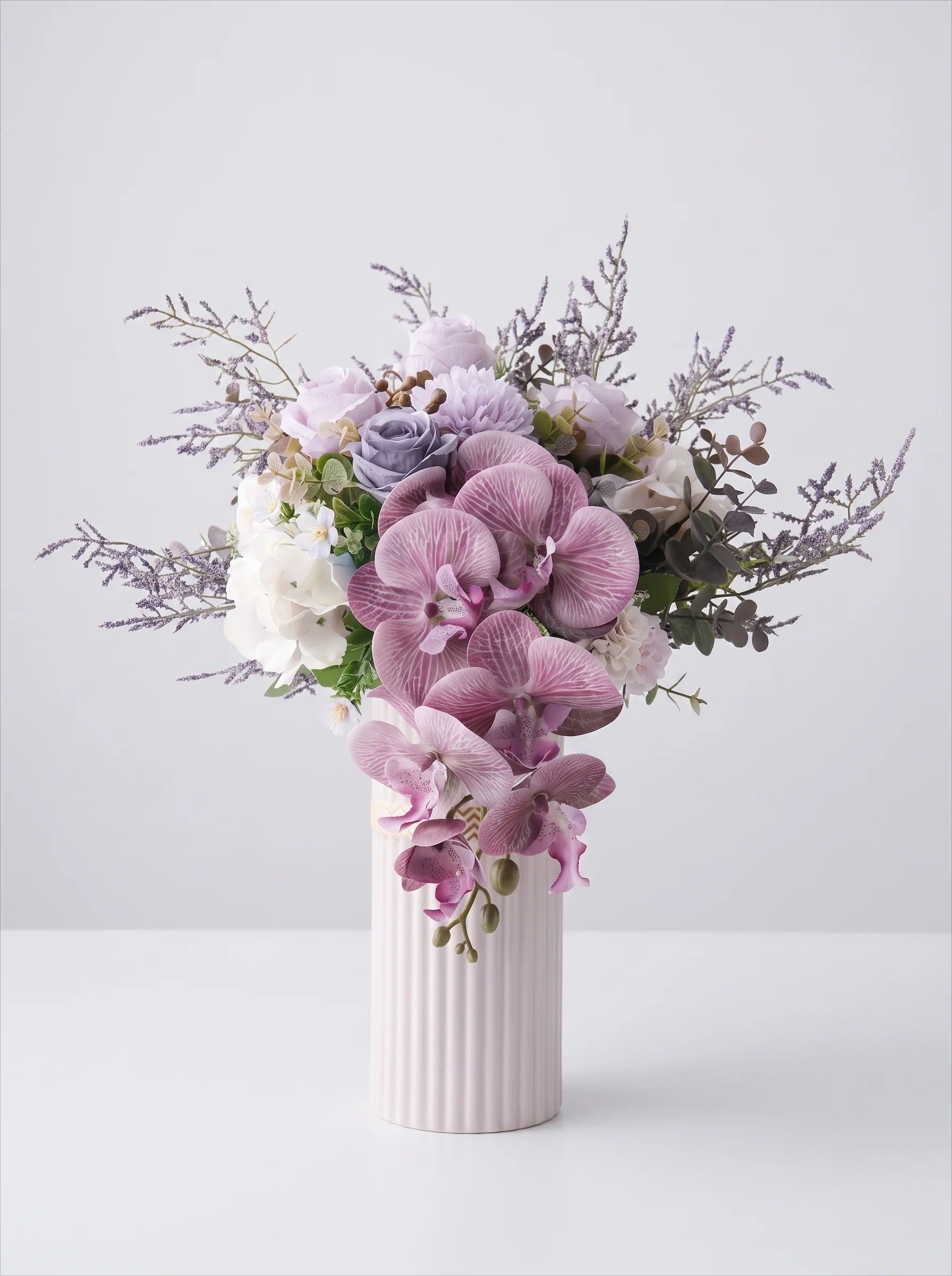 A floral arrangement featuring pink and purple orchids, lavender-toned roses, white blossoms, and green foliage in a ribbed white ceramic vase against a light gray studio background.