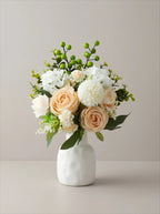 A floral arrangement in a white ceramic vase, featuring peach and white roses, white blossoms, green berries, and green foliage, set against a neutral beige background.