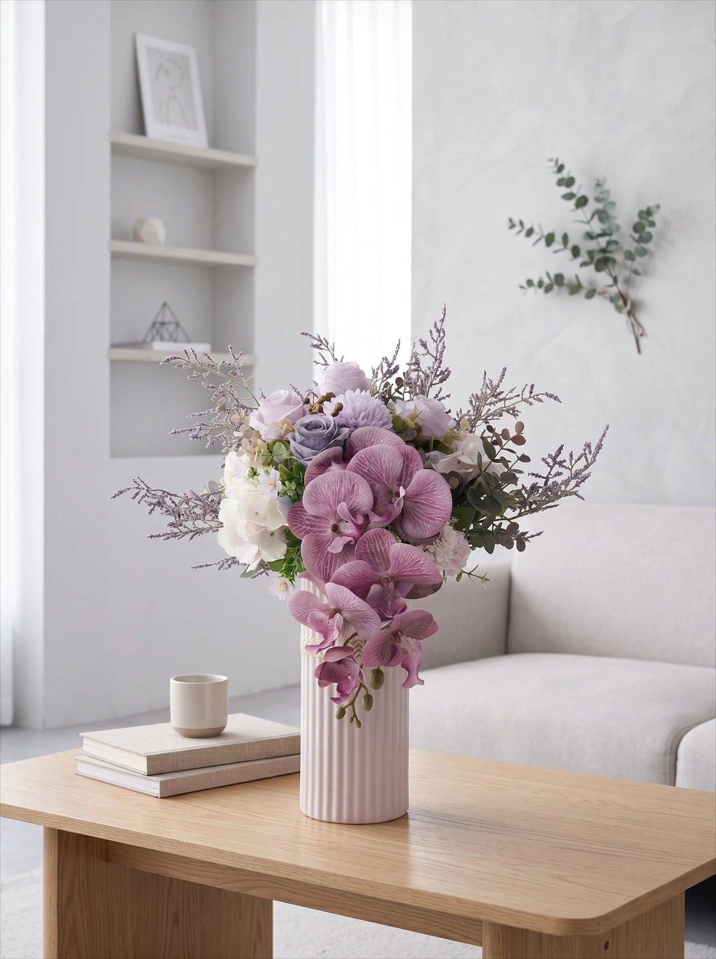 A pastel floral arrangement in a ribbed pale pink vase sits on a light wood table in a bright, modern living room with a sofa and shelves in the background.