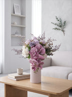 A pastel floral arrangement in a ribbed pale pink vase sits on a light wood table in a bright, modern living room with a sofa and shelves in the background.