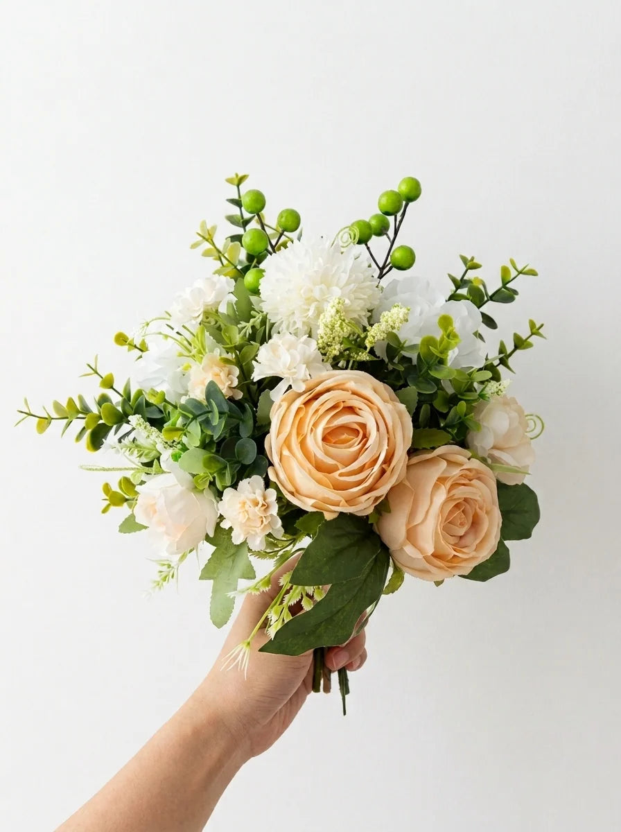 An artificial floral bouquet with peach roses, white flowers, green foliage, and green berries, held by a hand against a plain white background.