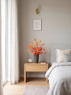 A styled bedroom corner featuring a light wood nightstand with a dark textured vase of orange autumn leaves, a small clock, and a book. The nightstand is positioned next to a bed with neutral gray and white bedding, under a brass wall sconce and a minimalist line-art print on a warm gray wall.