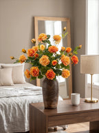 A large bouquet of orange and yellow roses in a dark ribbed ceramic vase, styled on a wooden bedside table in a modern, softly lit bedroom.