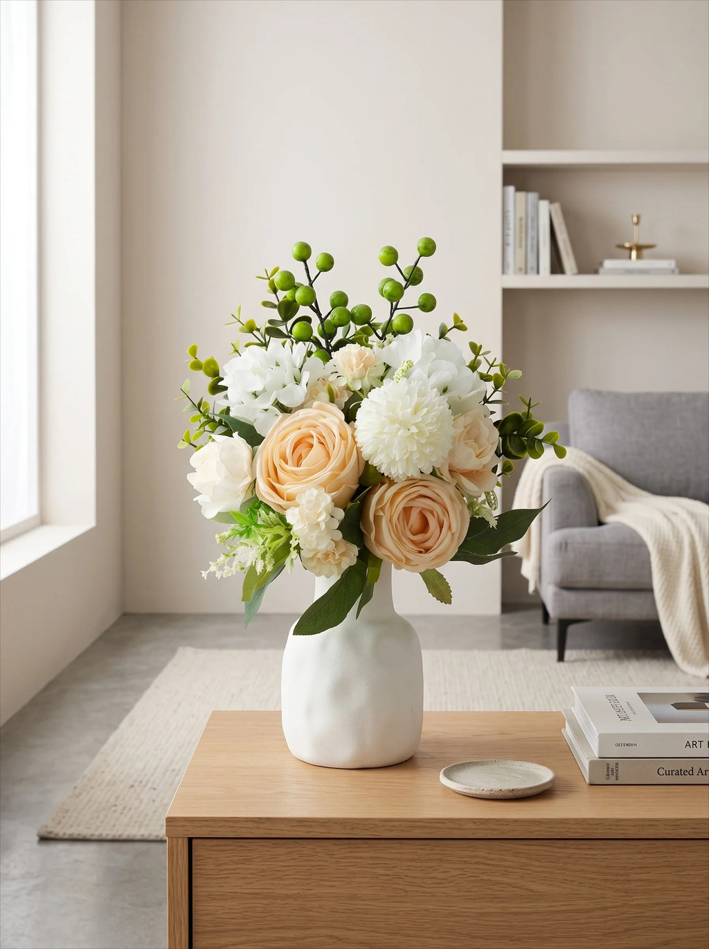 A white ceramic vase filled with a floral arrangement of peach roses, white blossoms, and green berries, placed on a wooden table in a bright, modern living room.