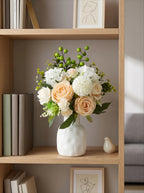 A white ceramic vase filled with a floral arrangement of peach roses, white flowers, and green berries, displayed on a light wood bookshelf in a cozy room setting.
