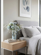 A styled bedroom scene featuring a light wood nightstand with a ribbed light blue vase holding a bouquet of pale blue and white flowers, positioned next to a bed with gray and white bedding.