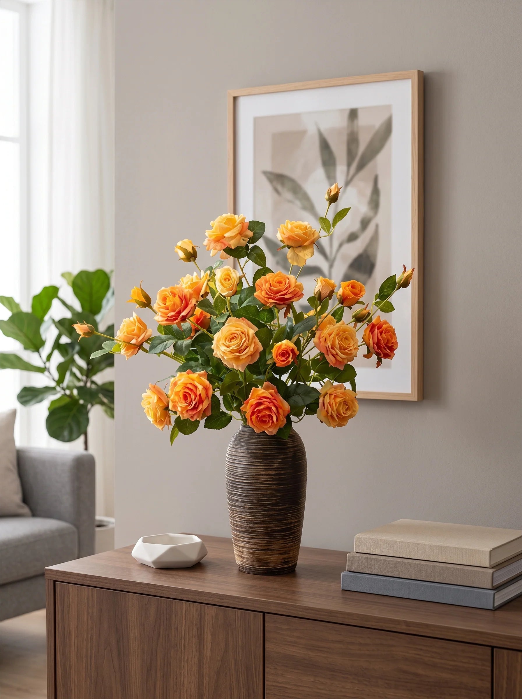 A vase filled with orange and yellow roses sits on a wooden console table in a styled interior room, with a framed botanical print on the wall, books, and a plant in the background.