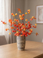 A faux autumn leaf arrangement in a textured gray and white striped ceramic vase, displayed on a light wood table in a neutral-toned room.