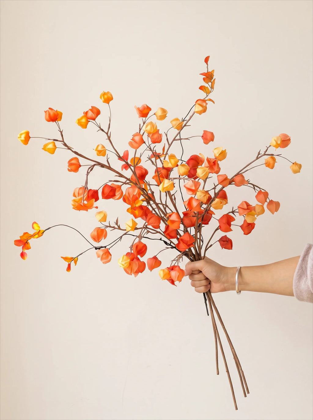A hand holding a bouquet of artificial autumn branches with orange, red, and yellow berries against a plain beige background.
