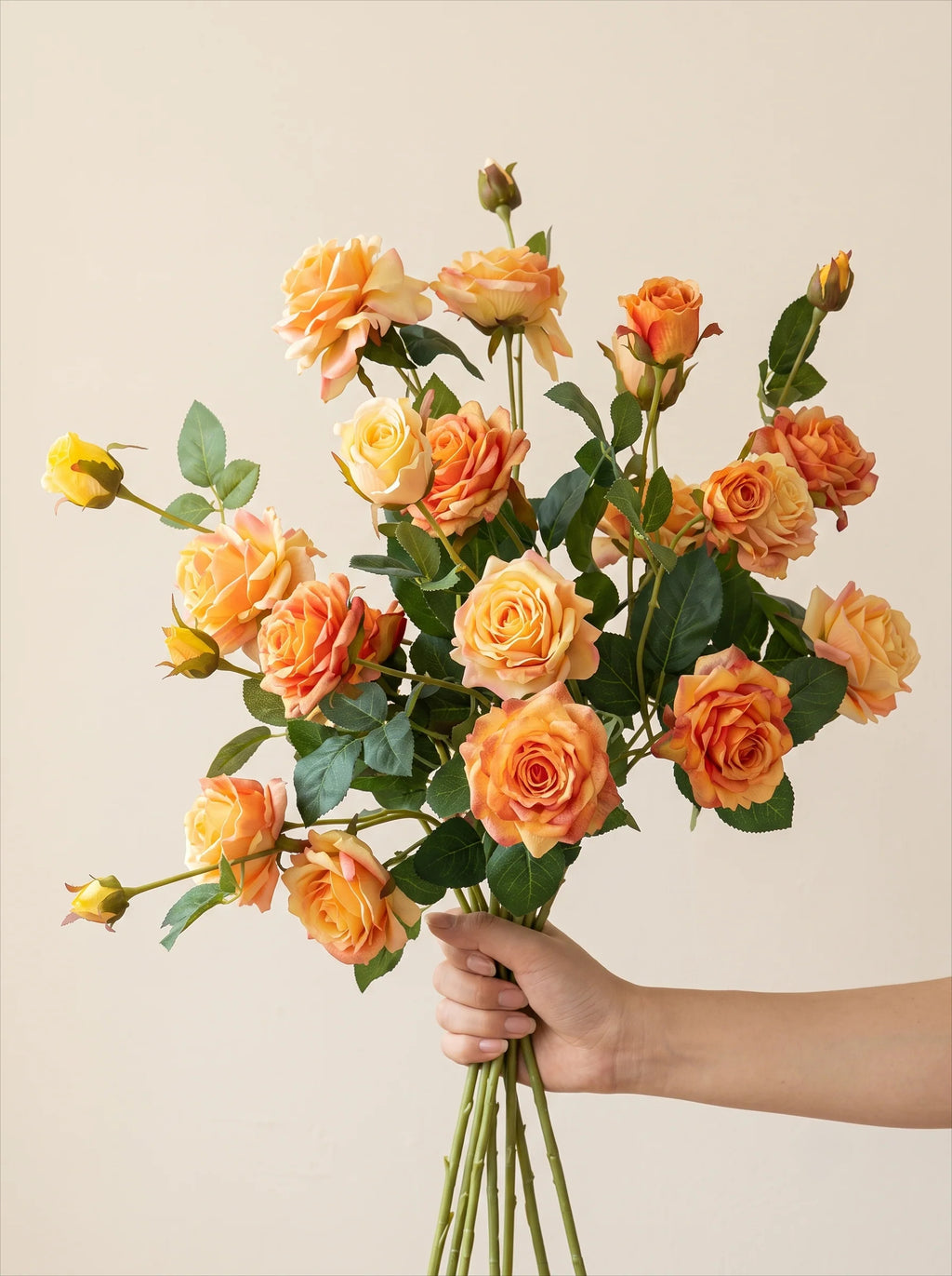 A bouquet of artificial orange and yellow roses with green stems and leaves, held by a person's hand against a plain beige background.
