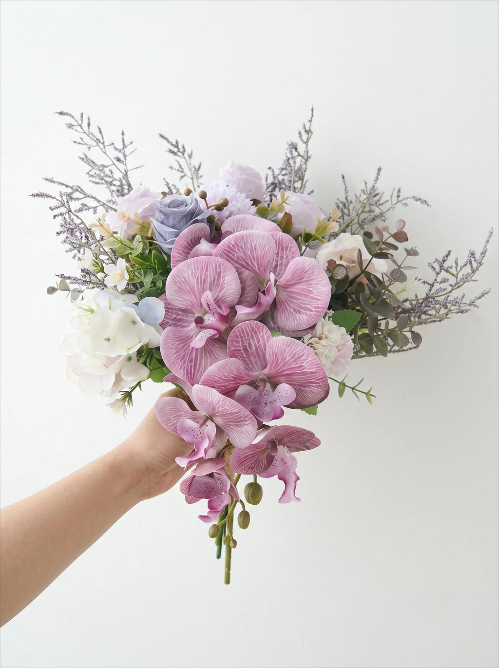 A hand holding a bouquet of artificial flowers, featuring prominent pink and purple orchids, white roses, lavender sprigs, and green foliage against a plain white background.