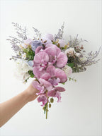 A hand holding a bouquet of artificial flowers, featuring prominent pink and purple orchids, white roses, lavender sprigs, and green foliage against a plain white background.