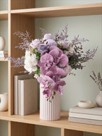 A floral arrangement of lavender orchids, white flowers, and purple filler stems in a pale pink ribbed vase, displayed on a wooden shelf with books and decor.