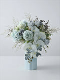 A floral arrangement in a light blue ribbed vase, featuring pale blue and white flowers with soft green foliage, photographed against a clean light gray studio background.
