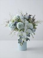 A floral arrangement in a light blue ribbed vase, featuring pale blue and white flowers with soft green foliage, photographed against a clean light gray studio background.