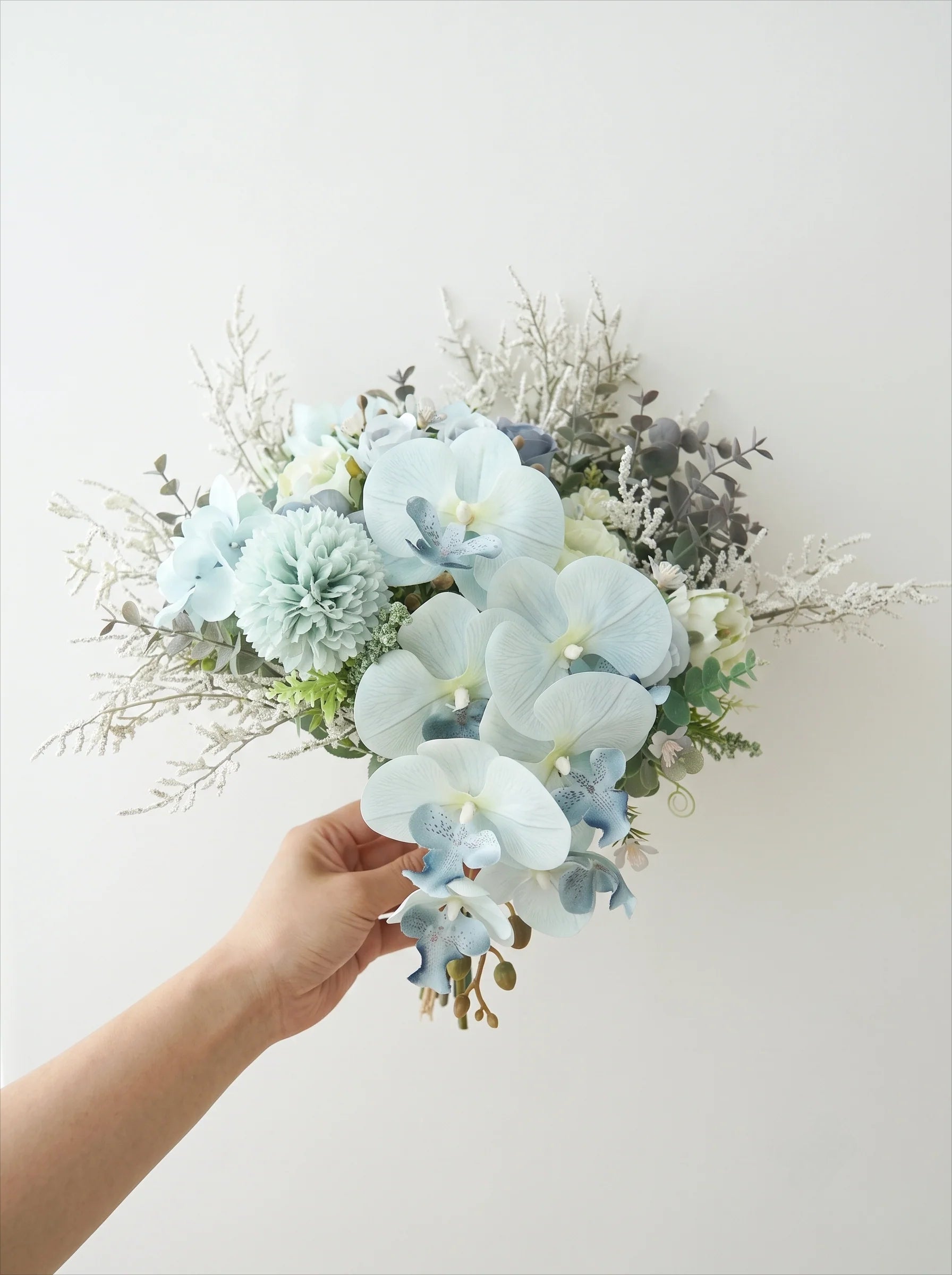 A hand holding a bouquet of artificial flowers, featuring light blue orchids, pale blue pompons, and muted gray-green foliage, set against a plain off-white background.
