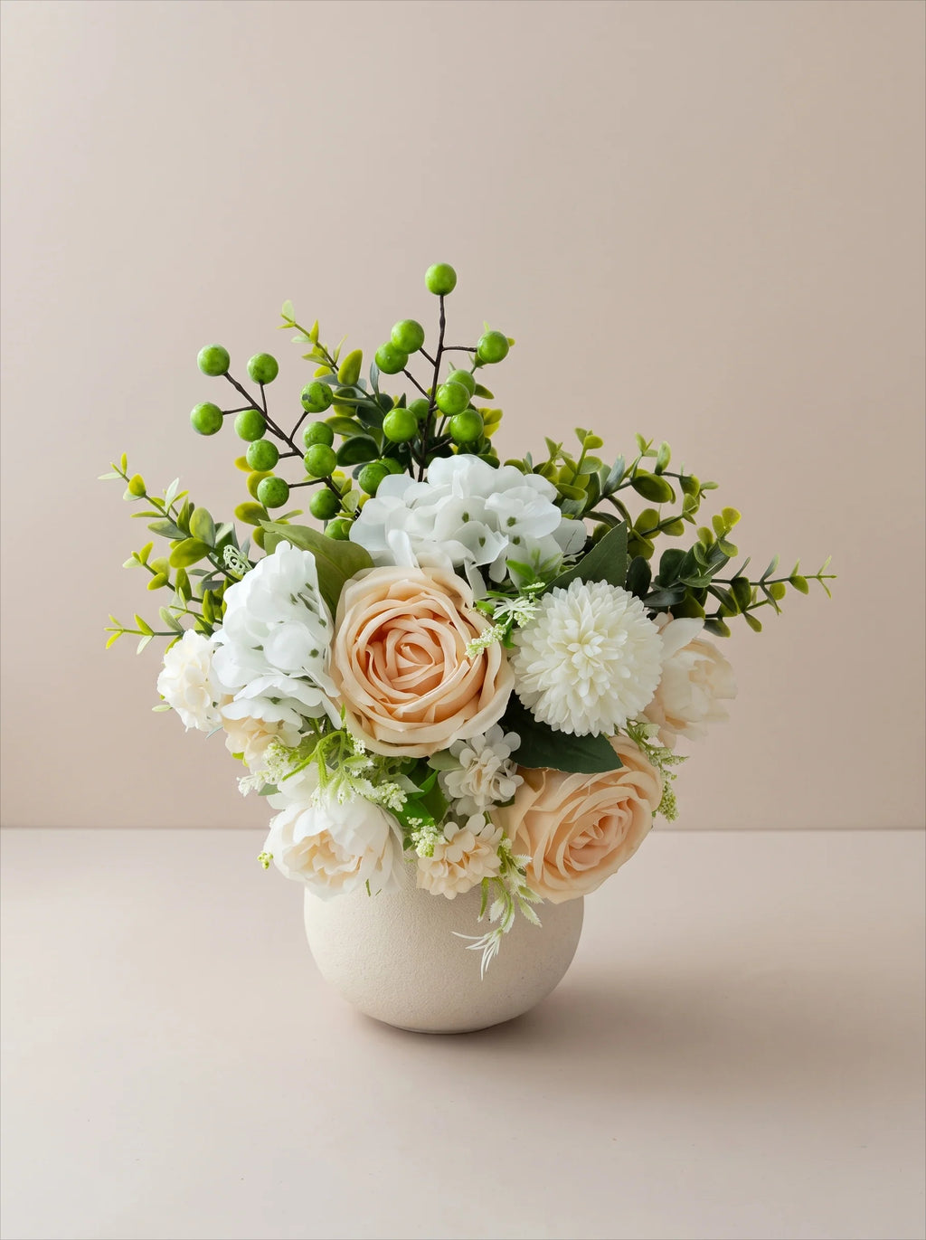 An artificial floral arrangement in a round white ceramic vase, featuring peach roses, white dahlia-like blooms, green berries, and green foliage, set against a solid beige background.