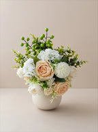 An artificial floral arrangement in a round white ceramic vase, featuring peach roses, white dahlia-like blooms, green berries, and green foliage, set against a solid beige background.