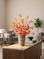 A large artificial floral arrangement with orange and yellow berries in a textured beige and brown striped vase, displayed on a wooden coffee table in a modern living room setting.