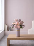 A pastel pink and lavender floral arrangement in a ribbed lavender vase with a gold band, displayed on a light wood table in a softly lit modern room.