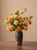 A bouquet of orange and yellow roses with green leaves arranged in a dark brown textured ceramic vase, set on a wooden table against a solid beige background.