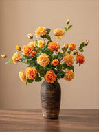 A bouquet of orange and yellow roses with green leaves arranged in a dark brown textured ceramic vase, set on a wooden table against a solid beige background.